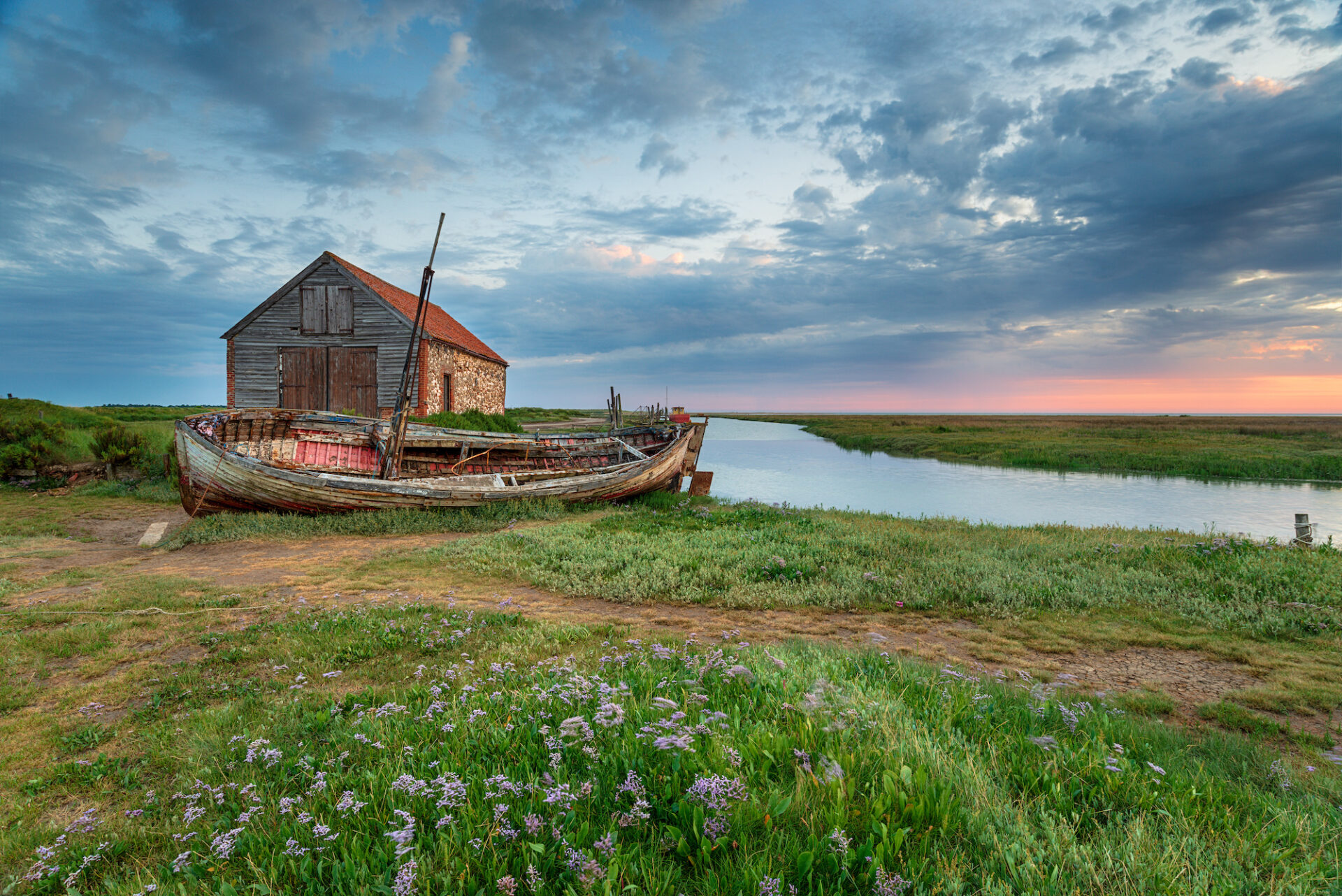 A grassy field with a barn and a boat and morning sky in the background